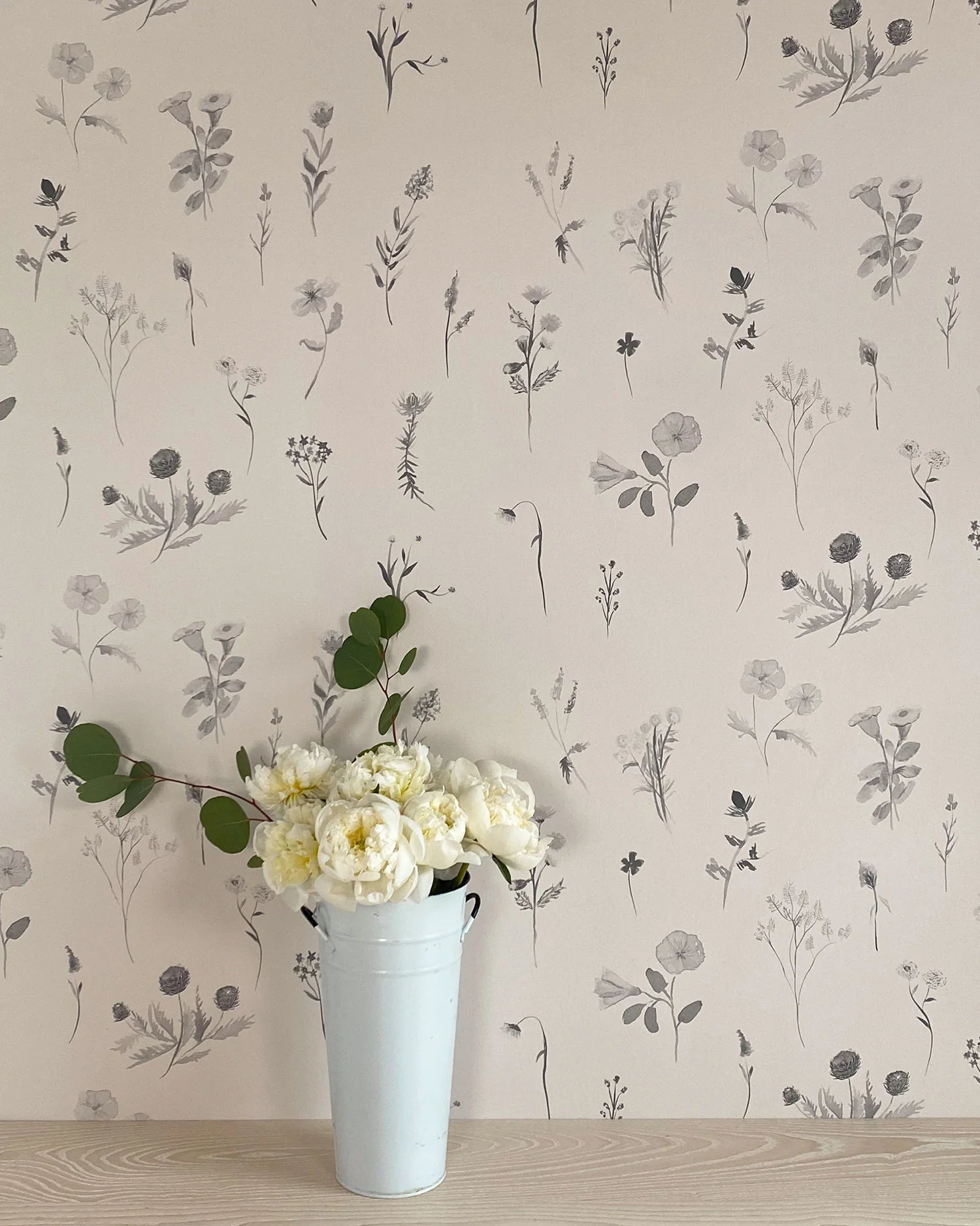 A vase of flowers stands in front of a wall covered in a painterly flower print in shades of gray on a cream field.