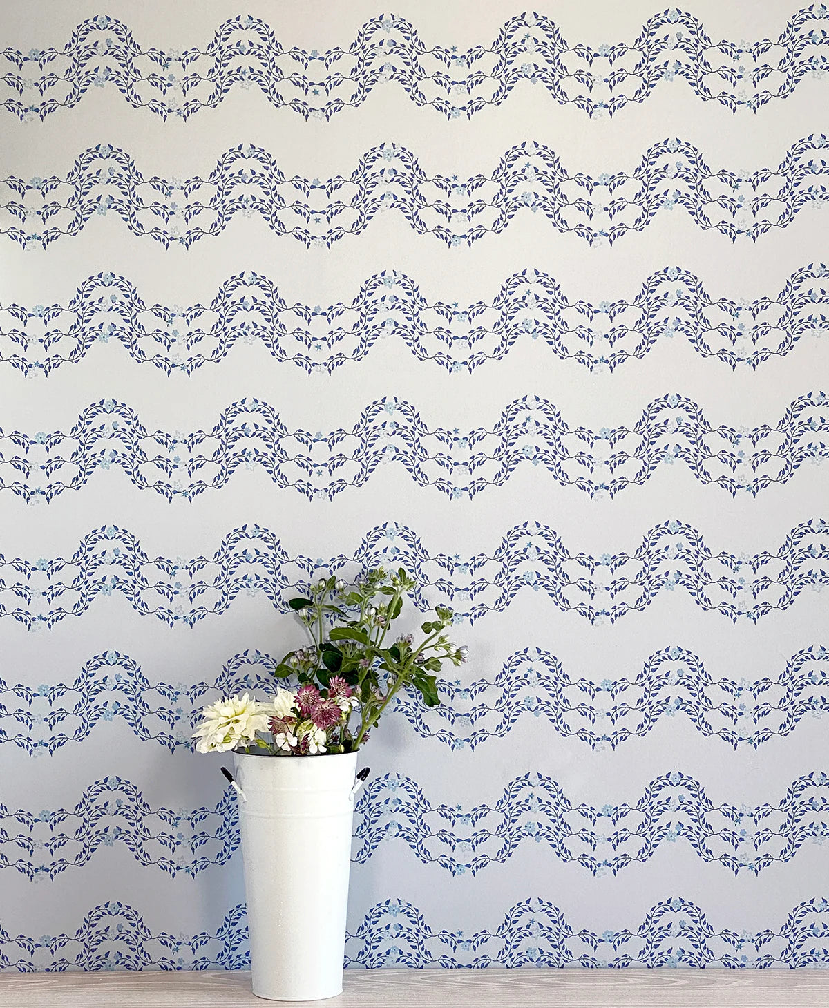 A vase of flowers stands in front of a wall covered in a curving floral stripe print in shades of blue on a cream field.