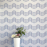 A vase of flowers stands in front of a wall covered in a curving floral stripe print in shades of blue on a cream field.