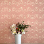 A vase of flowers stands in front of a wall covered in a curving floral stripe print in shades of cream on a pink field.