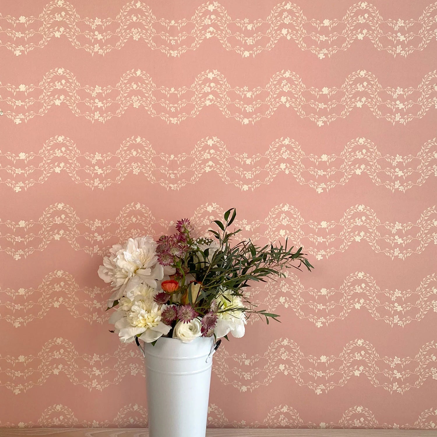 A vase of flowers stands in front of a wall covered in a curving floral stripe print in shades of cream on a pink field.