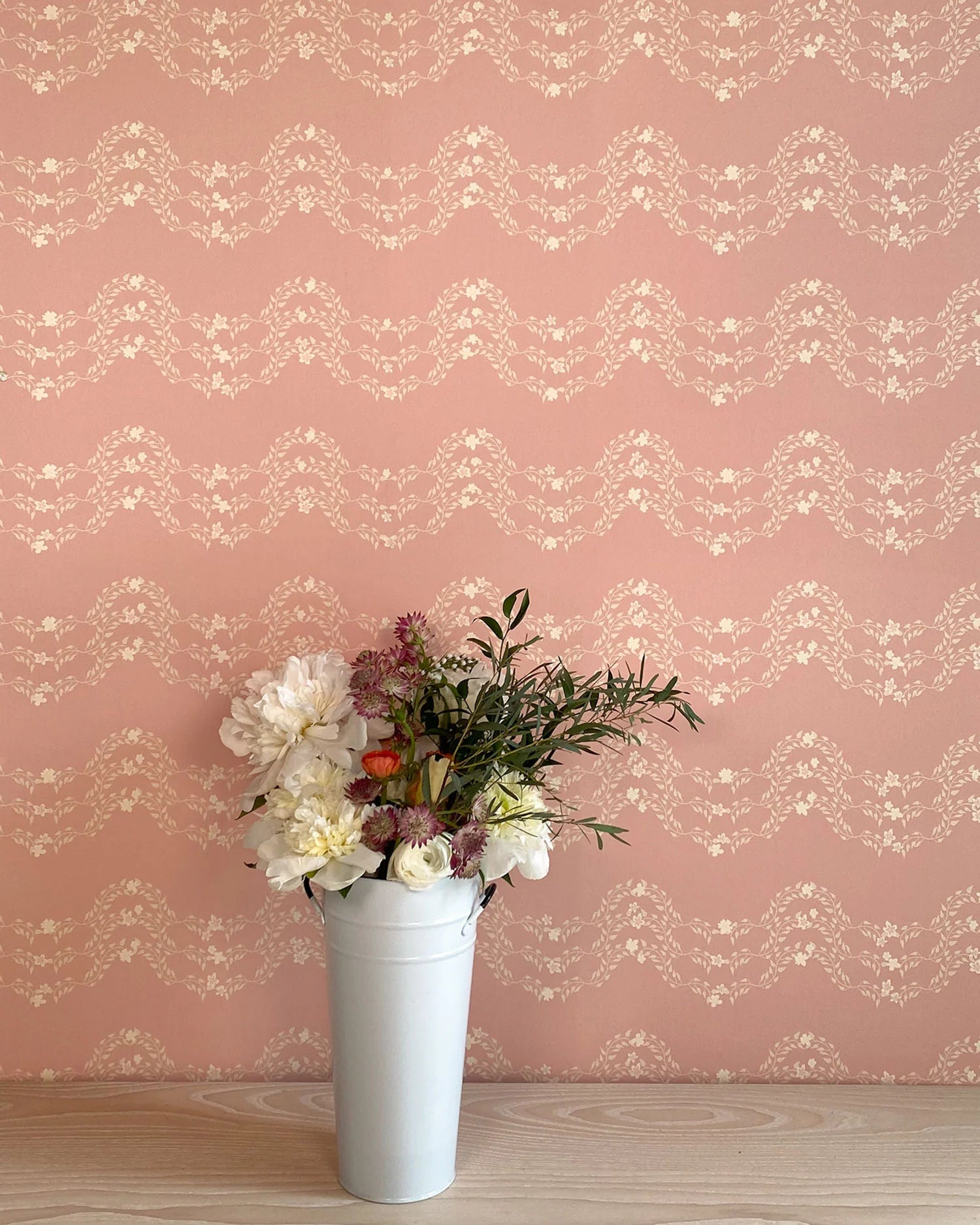 A vase of flowers stands in front of a wall covered in a curving floral stripe print in shades of cream on a pink field.