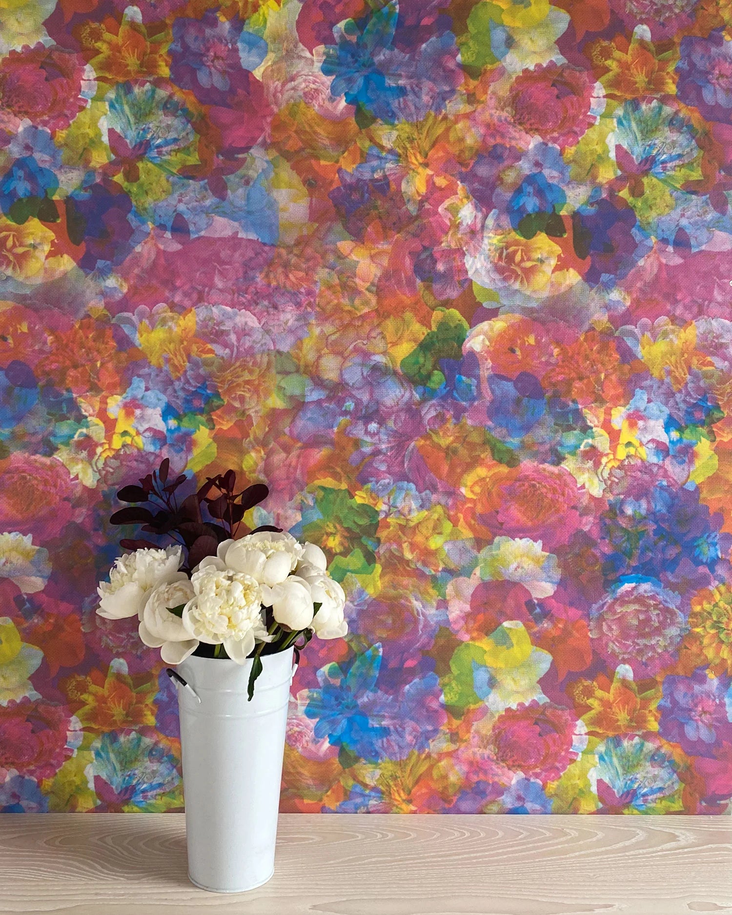 A vase of flowers stands in front of a wall covered in a dense photorealistic floral print in multicolor neon shades.