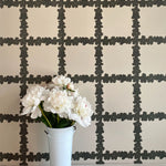 A vase of flowers stands in front of a wall covered in a floral grid print in brown on a white field.