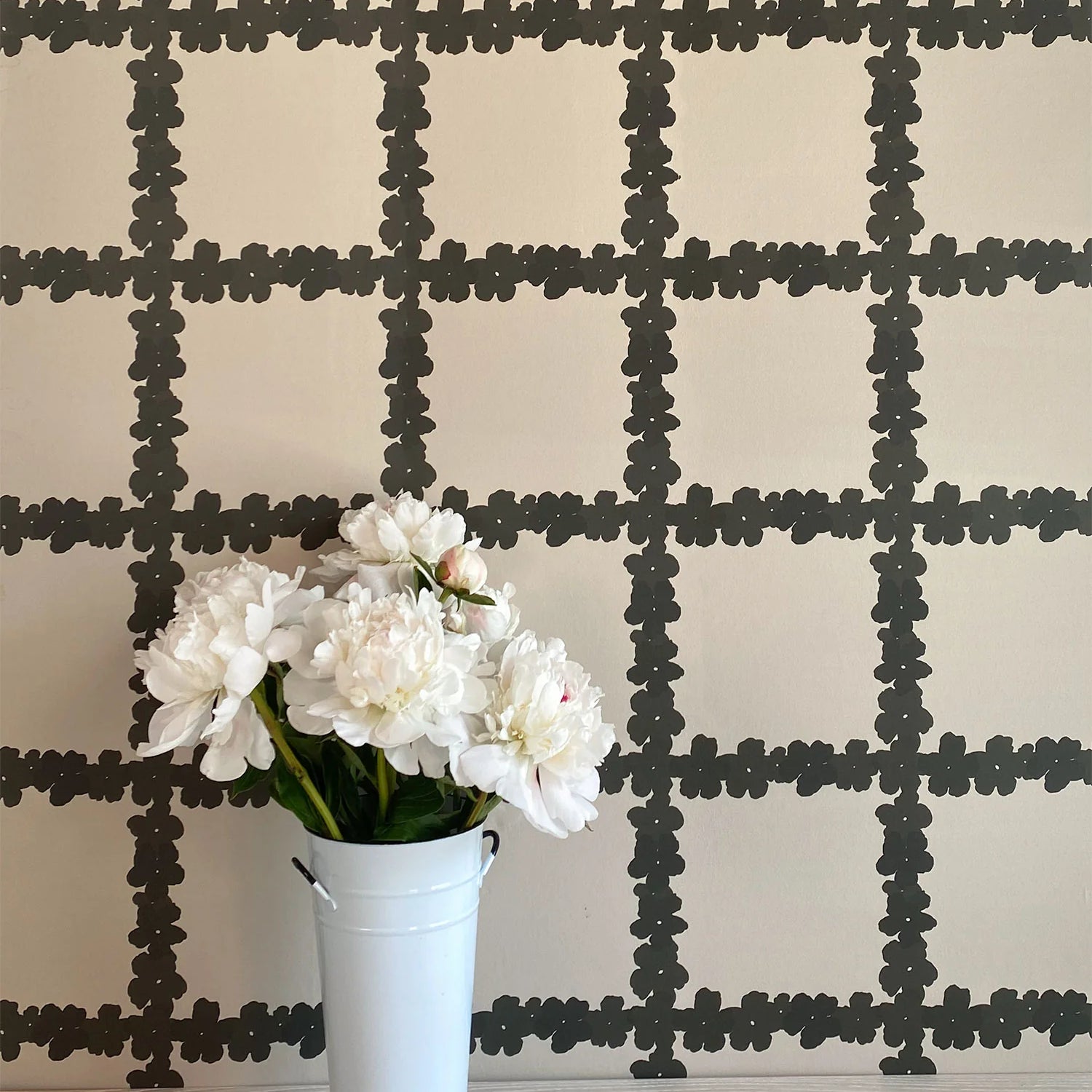 A vase of flowers stands in front of a wall covered in a floral grid print in brown on a white field.