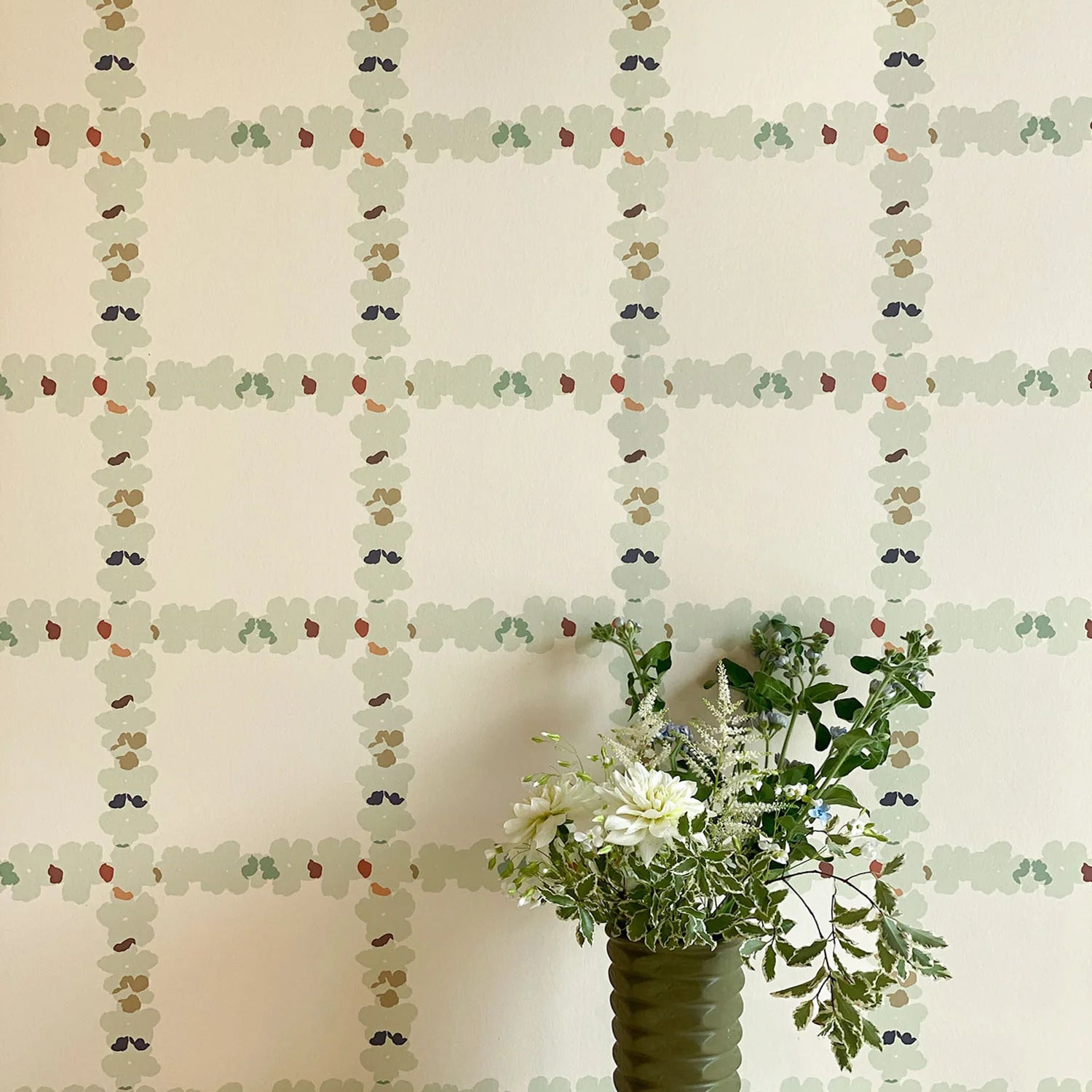 A vase of flowers stands in front of a wall covered in a floral grid print in cream, green and pink on a white field.