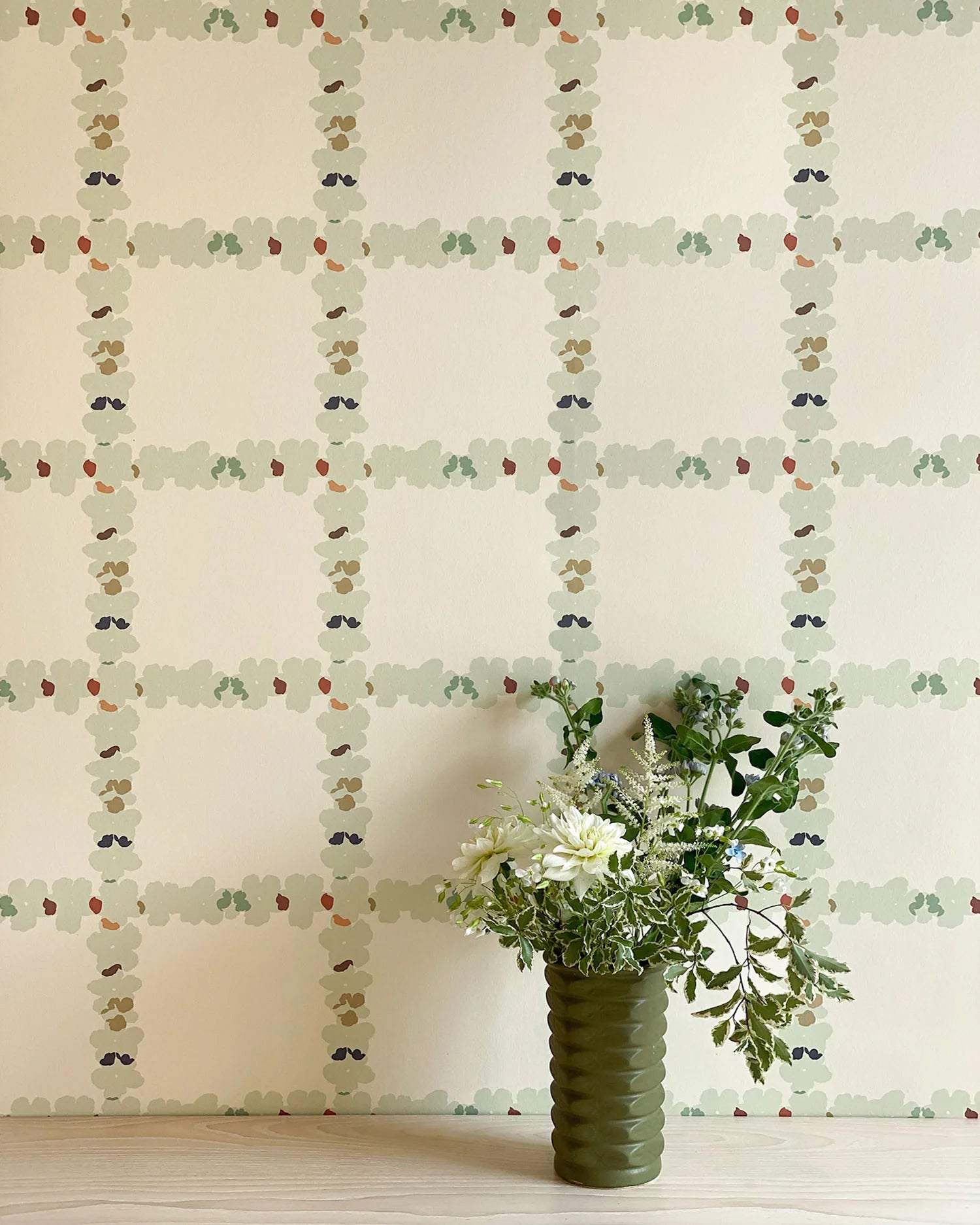 A vase of flowers stands in front of a wall covered in a floral grid print in cream, green and pink on a white field.