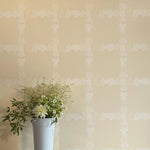 A vase of flowers stands in front of a wall covered in a floral grid print in cream on a tan field.