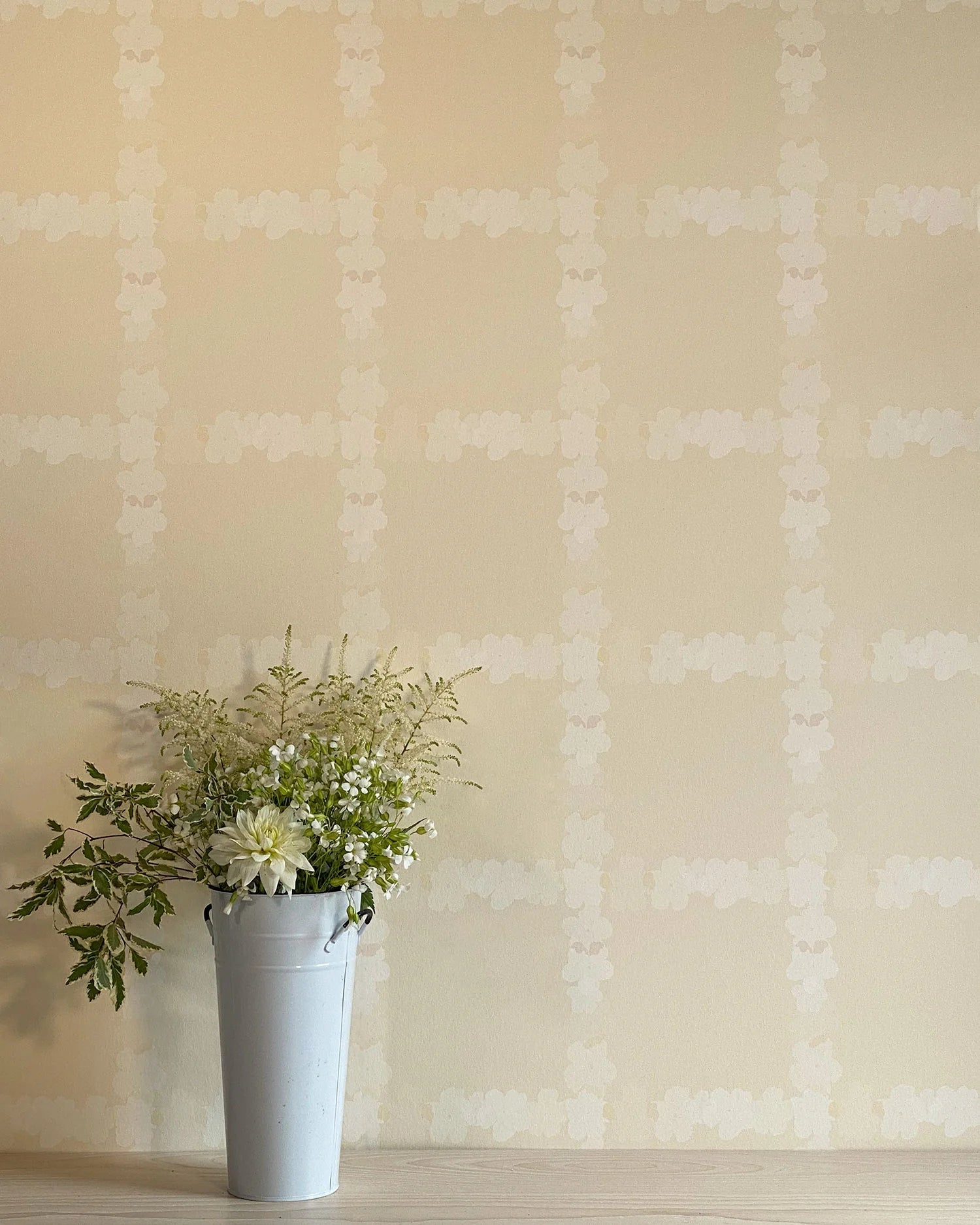 A vase of flowers stands in front of a wall covered in a floral grid print in cream on a tan field.