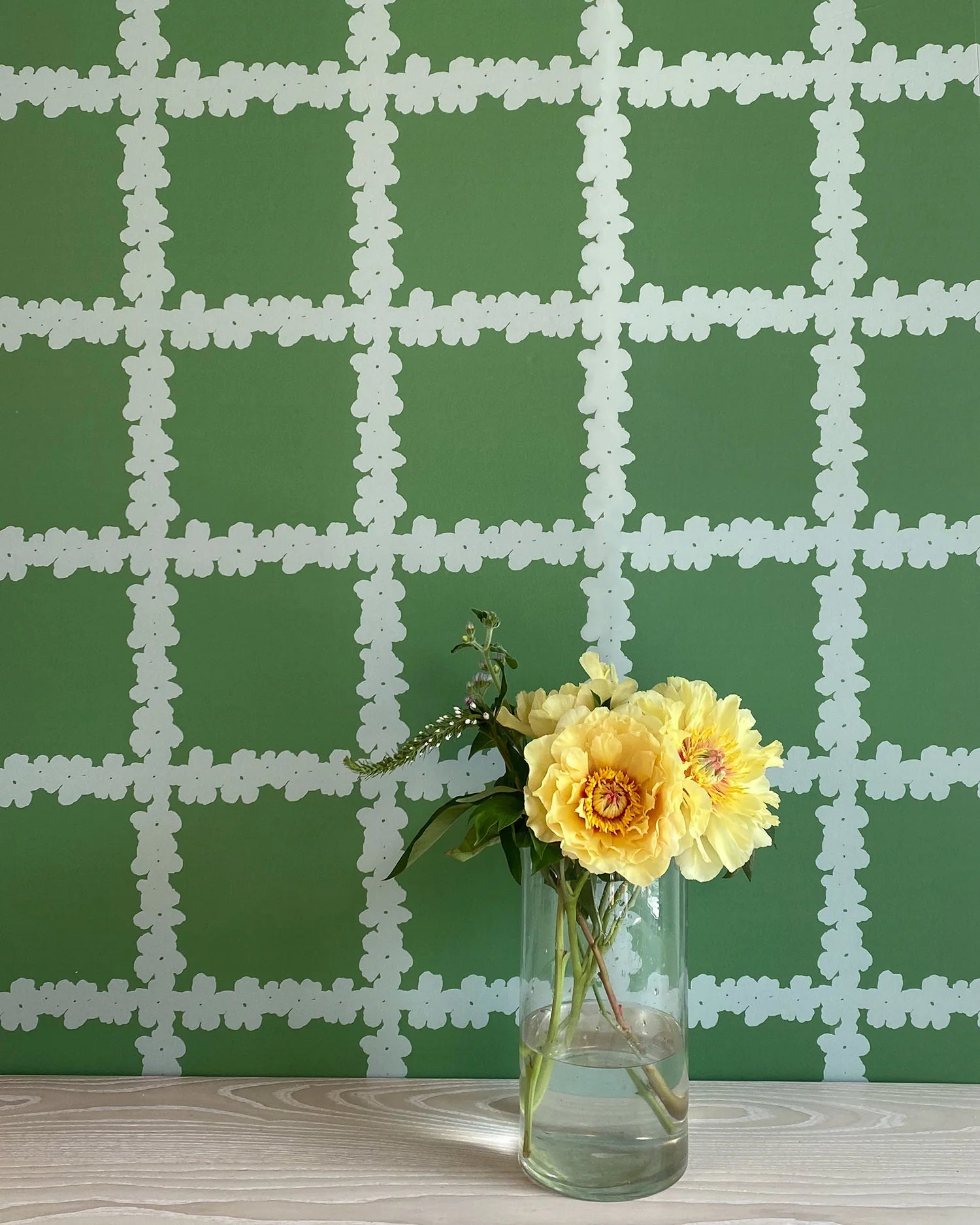 A vase of flowers stands in front of a wall covered in a floral grid print in white on a green field.