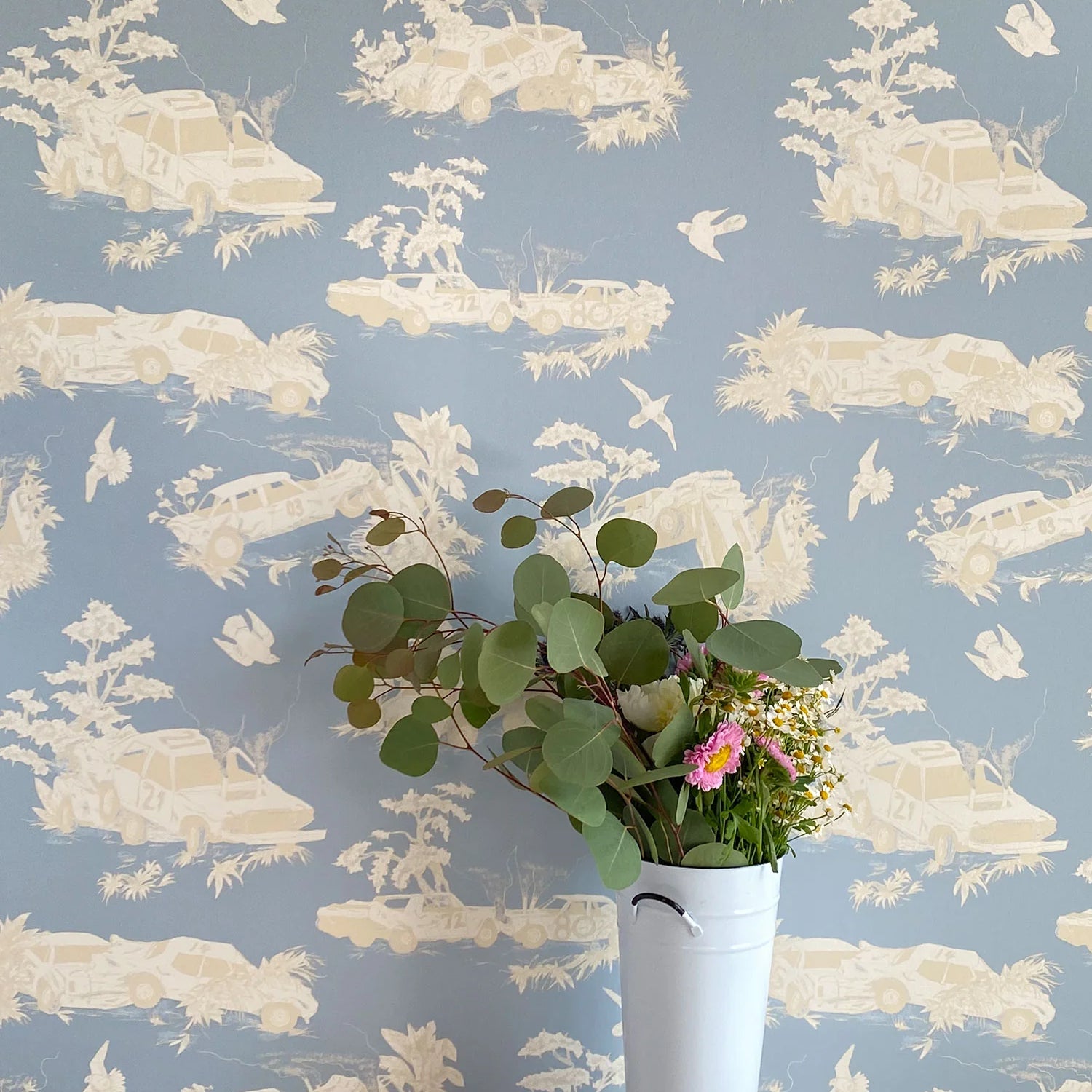 A vase of flowers stands in front of a wall covered in a playful illustrated car crash print in cream, white and light blue.