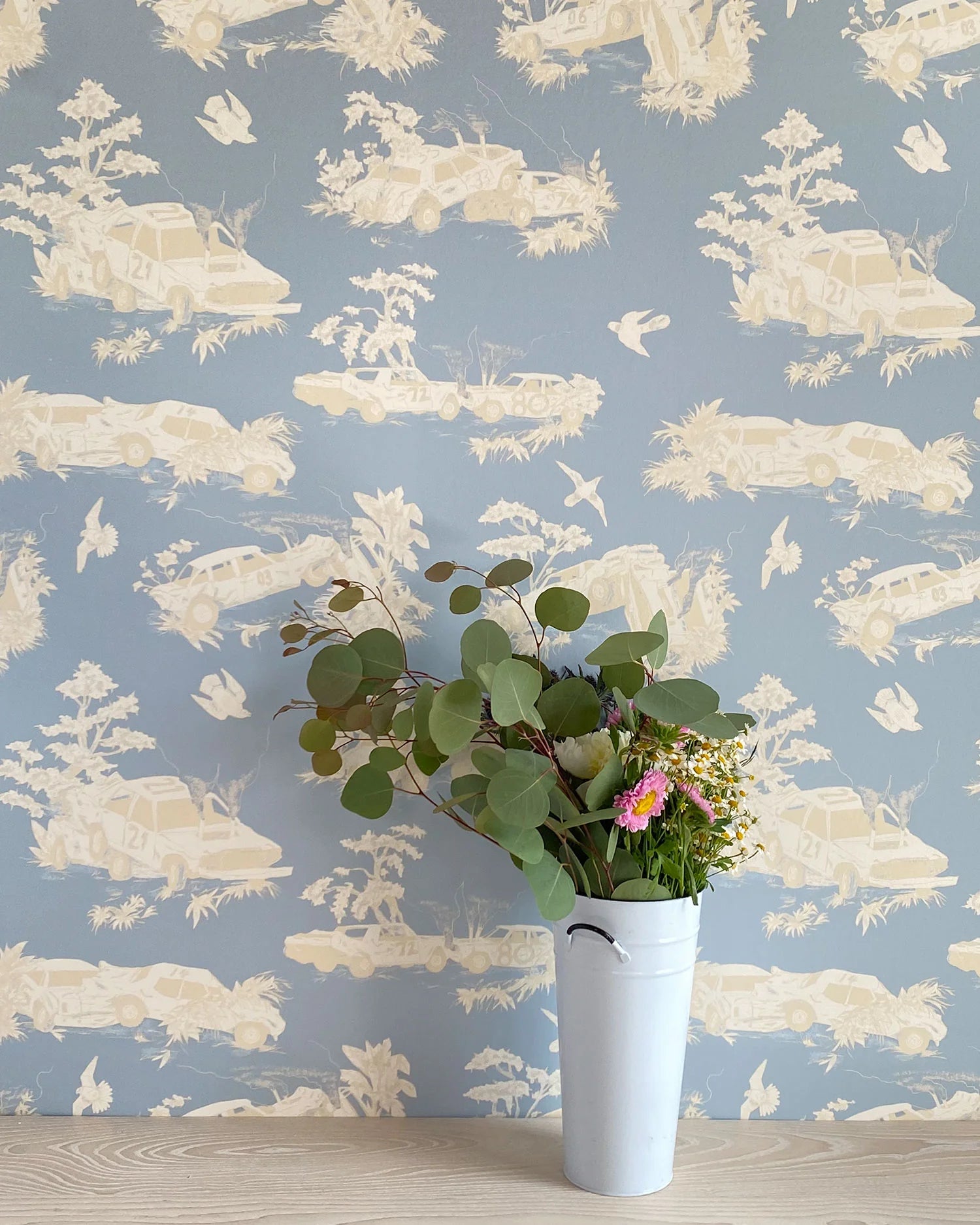 A vase of flowers stands in front of a wall covered in a playful illustrated car crash print in cream, white and light blue.