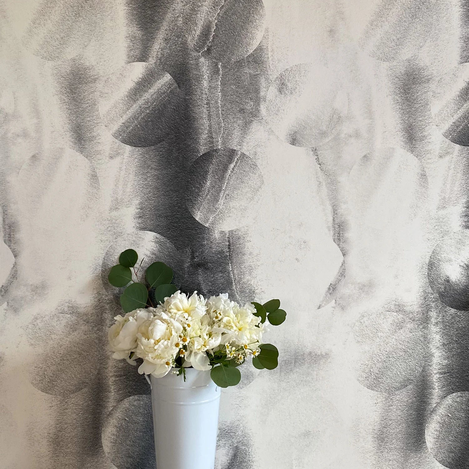 A vase of flowers stands in front of a wall papered in an abstract curvilinear pattern in mottled black and white.