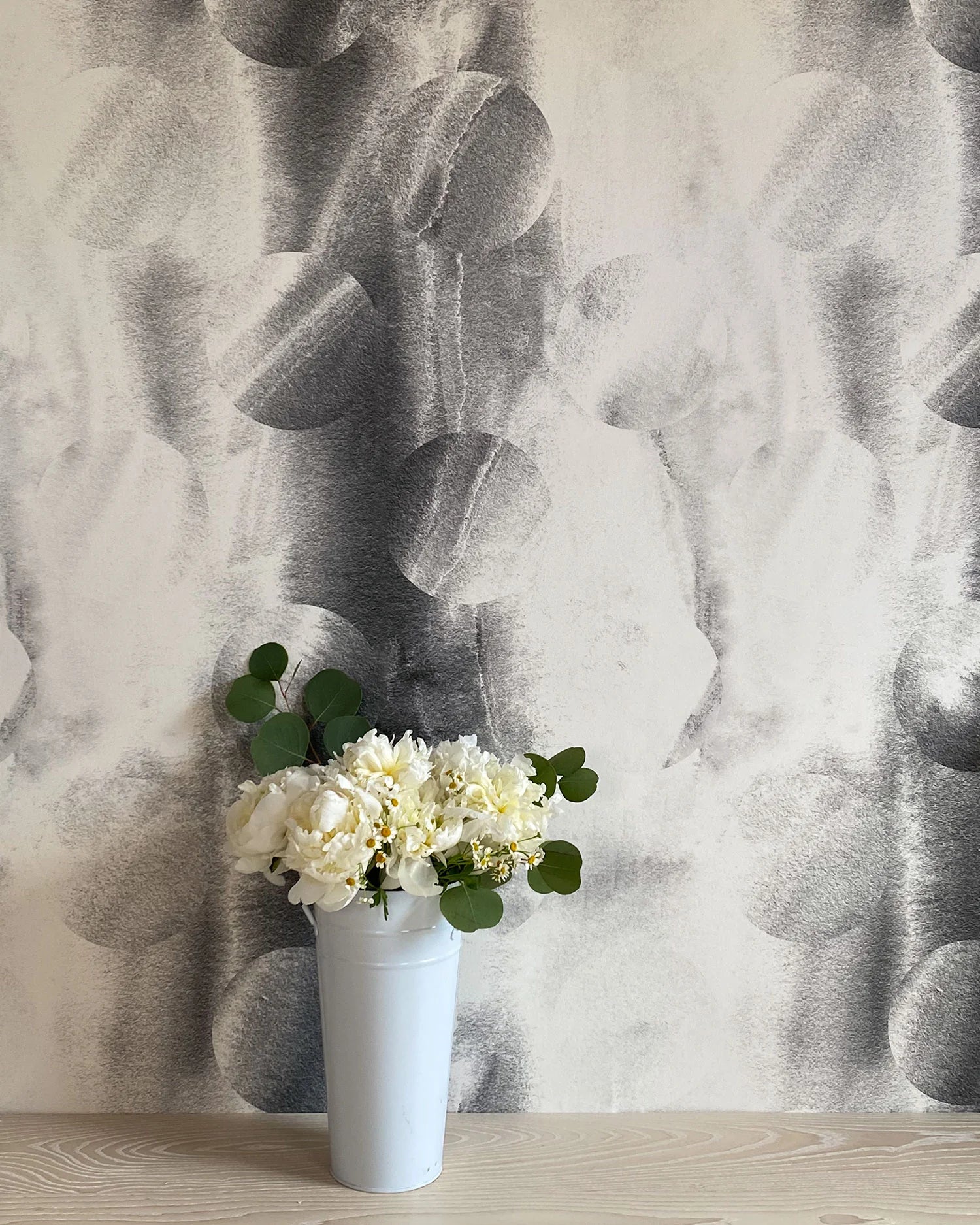 A vase of flowers stands in front of a wall papered in an abstract curvilinear pattern in mottled black and white.