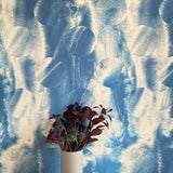 A vase of flowers stands in front of a wall papered in an abstract curvilinear pattern in mottled blue and tan.