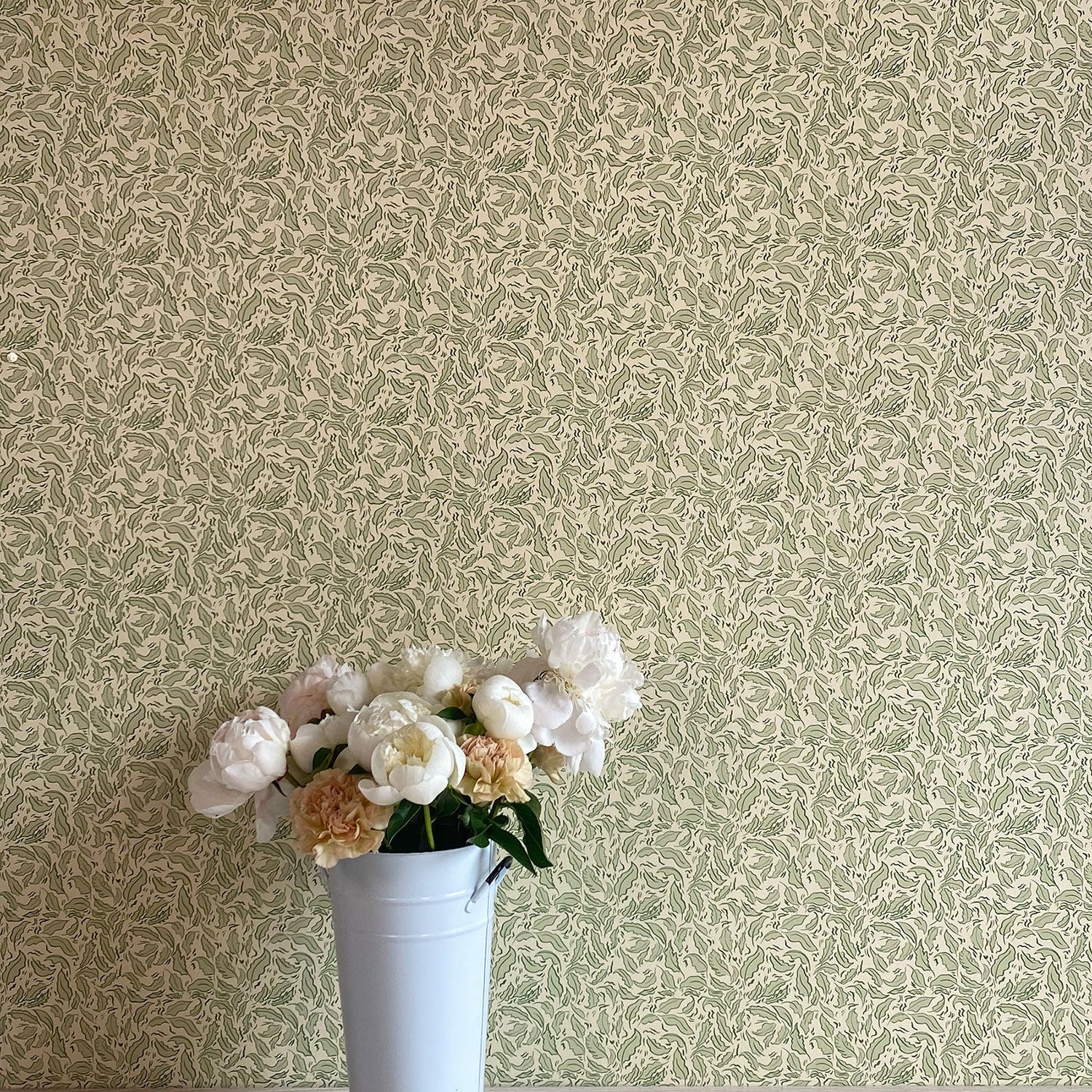 A vase of flowers stands in front of a wall papered in a painterly leaf print in olive and black on a cream field.