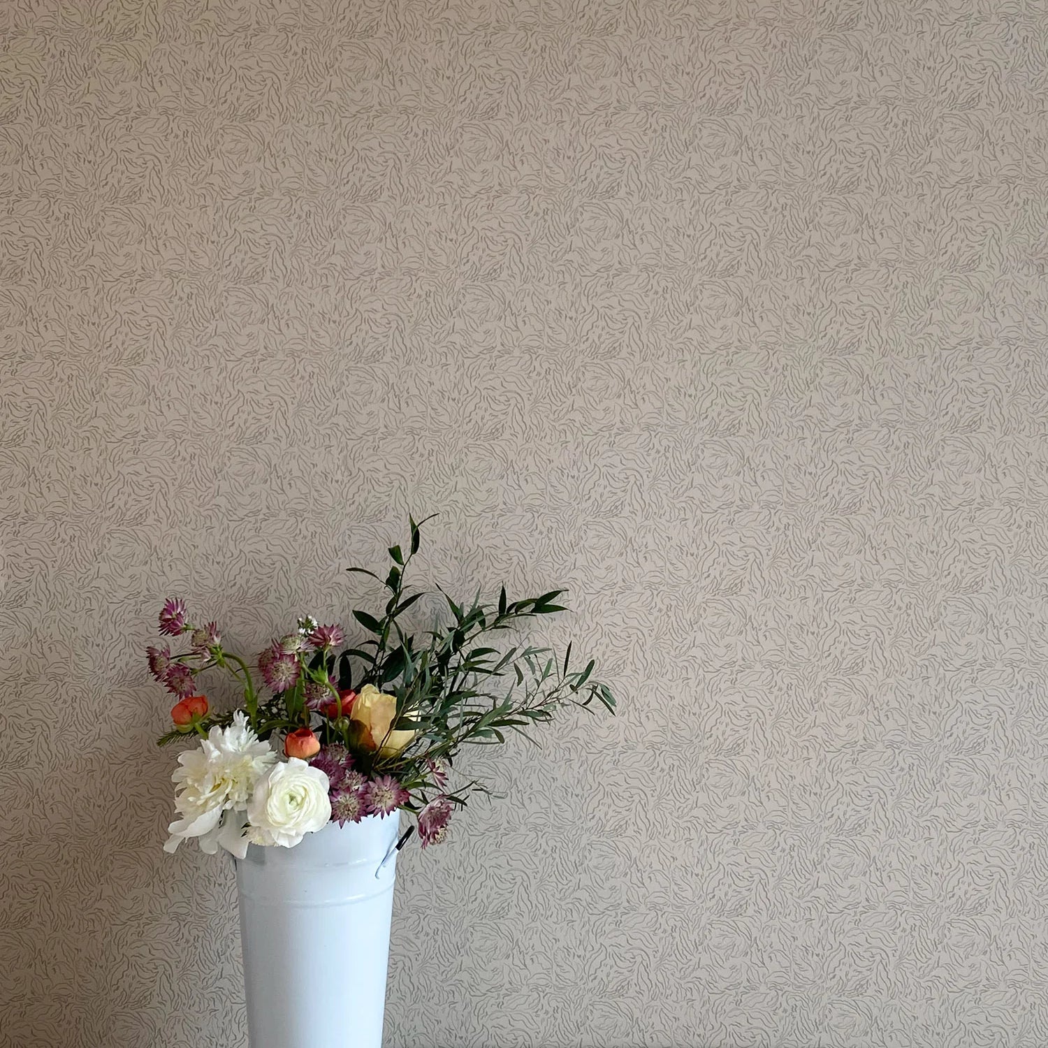 A vase of flowers stands in front of a wall papered in a painterly leaf print in black on a greige field.