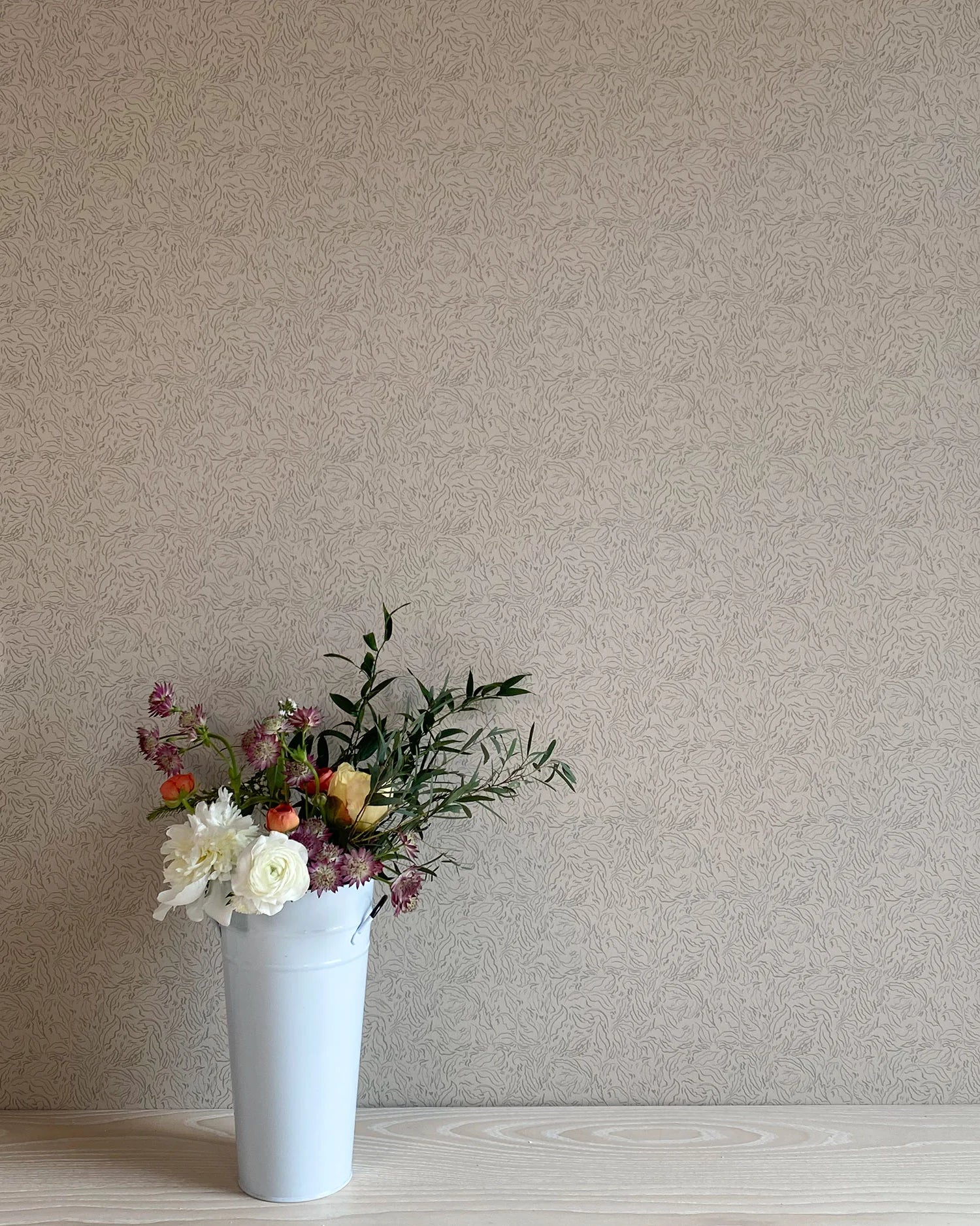 A vase of flowers stands in front of a wall papered in a painterly leaf print in black on a greige field.