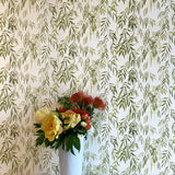 A vase of flowers stands in front of a wall papered in a painterly willow leaf print in green on a white field.