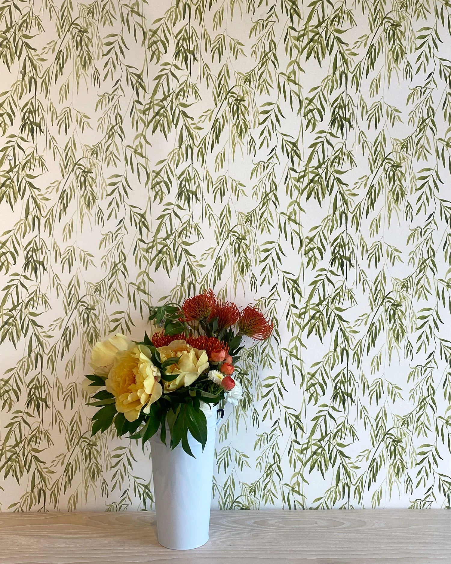 A vase of flowers stands in front of a wall papered in a painterly willow leaf print in green on a white field.