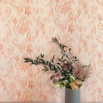 A vase of flowers stands in front of a wall papered in a painterly willow leaf print in coral on a cream field.