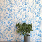 A vase of flowers stands in front of a wall papered in a painterly willow leaf print in blue on a white field.