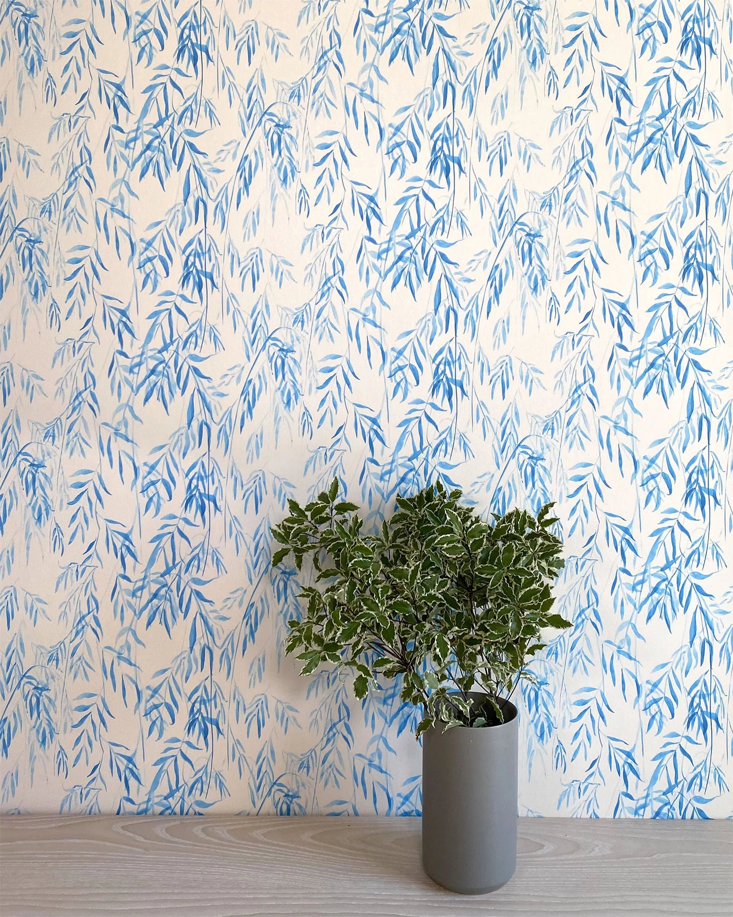 A vase of flowers stands in front of a wall papered in a painterly willow leaf print in blue on a white field.