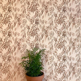 A potted plant stands in front of a wall papered in a painterly willow leaf print in brown on a cream field.