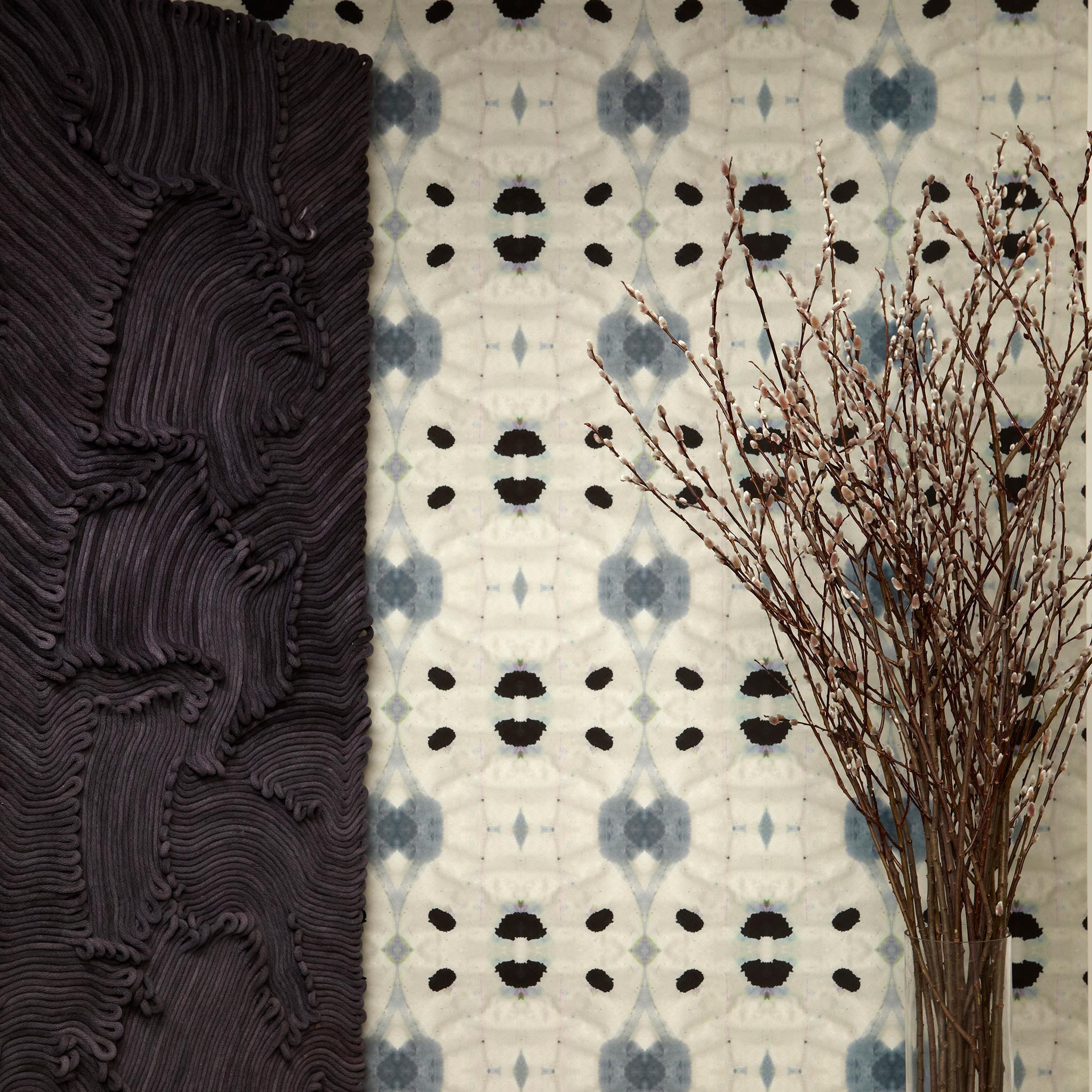 Flowers and a black room divider stand in front of a wall papered in an abstract ink blot print in charcoal, black and tan.