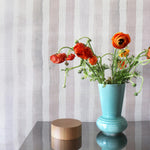 An end table with flowers stands in front of a wall papered in a painterly stripe print in pink, gray, cream and white.