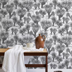 A bench with a towel and clay vases stands in front of a wall papered in a painterly tree print in charcoal and white.