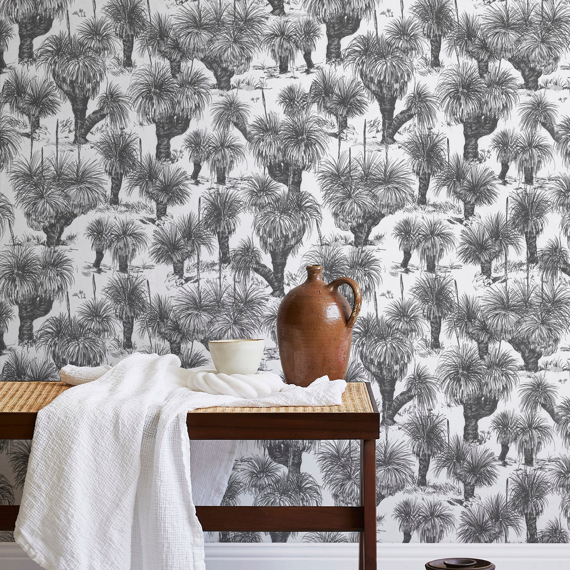 A bench with a towel and clay vases stands in front of a wall papered in a painterly tree print in charcoal and white.