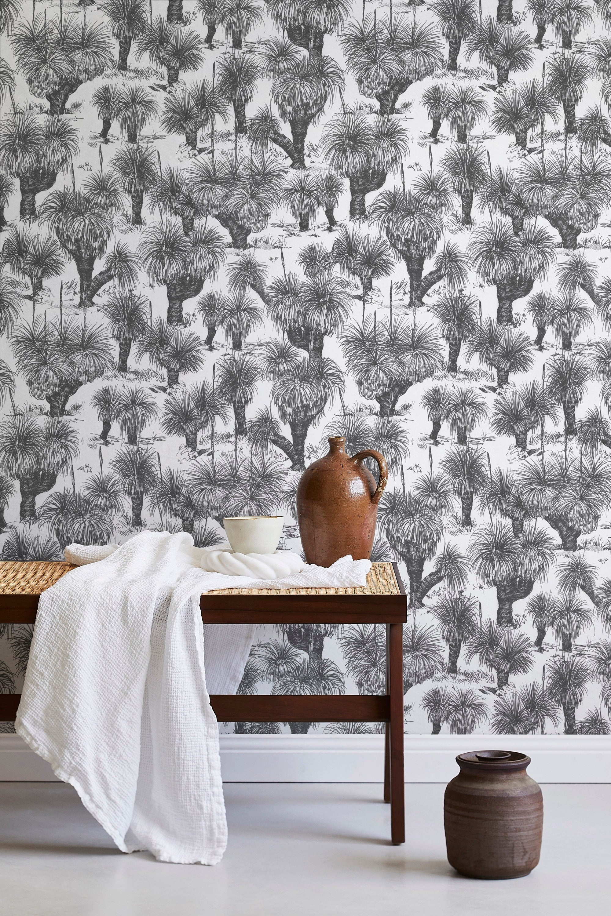 A bench with a towel and clay vases stands in front of a wall papered in a painterly tree print in charcoal and white.