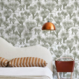 A modernist bed, hanging lamp and chair stand in front of a wall papered in a painterly tree print in sage and white.