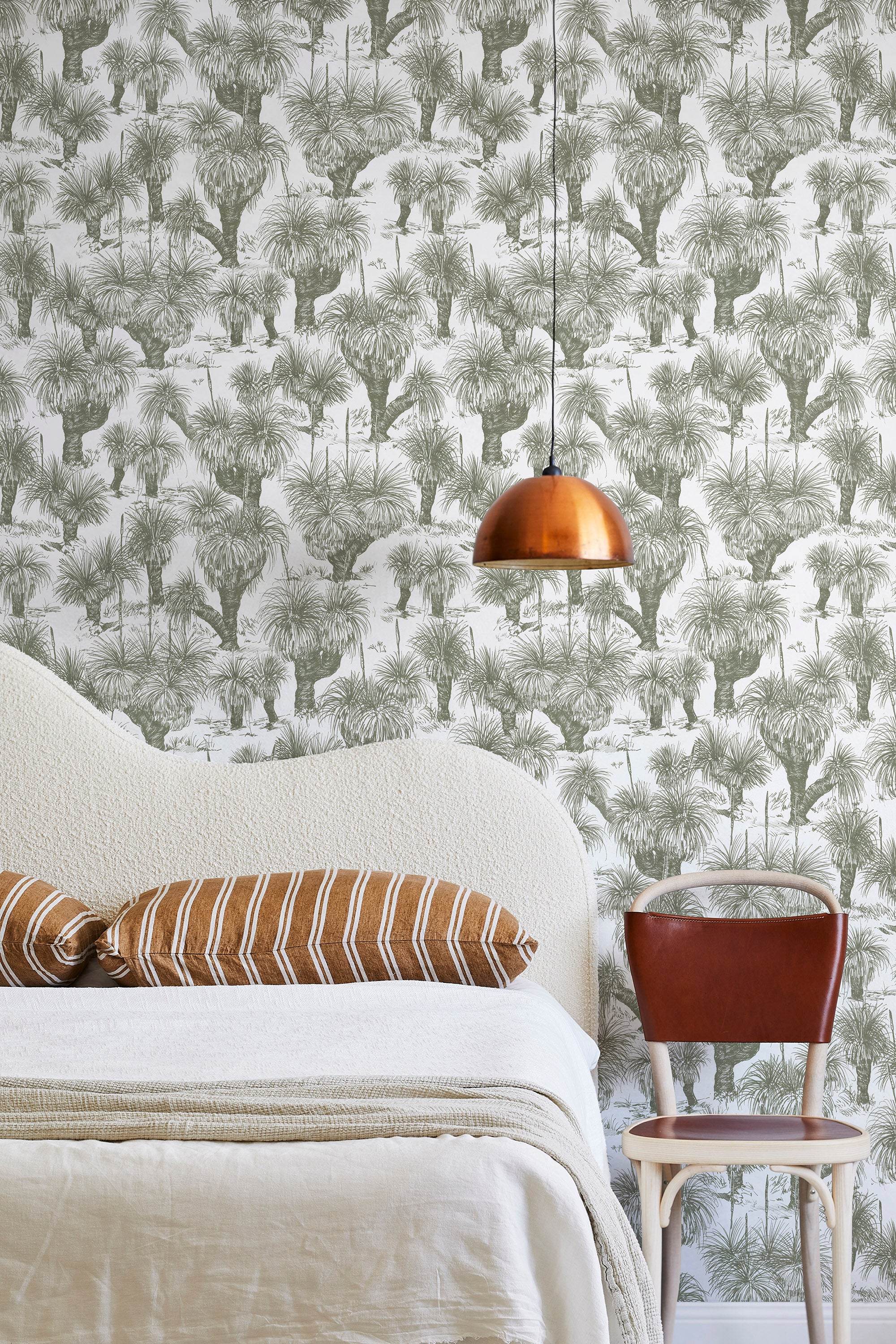 A modernist bed, hanging lamp and chair stand in front of a wall papered in a painterly tree print in sage and white.