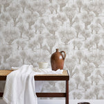 A bench with a towel and clay vases stands in front of a wall papered in a painterly tree print in tan and white.