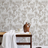 A bench with a towel and clay vases stands in front of a wall papered in a painterly tree print in tan and white.