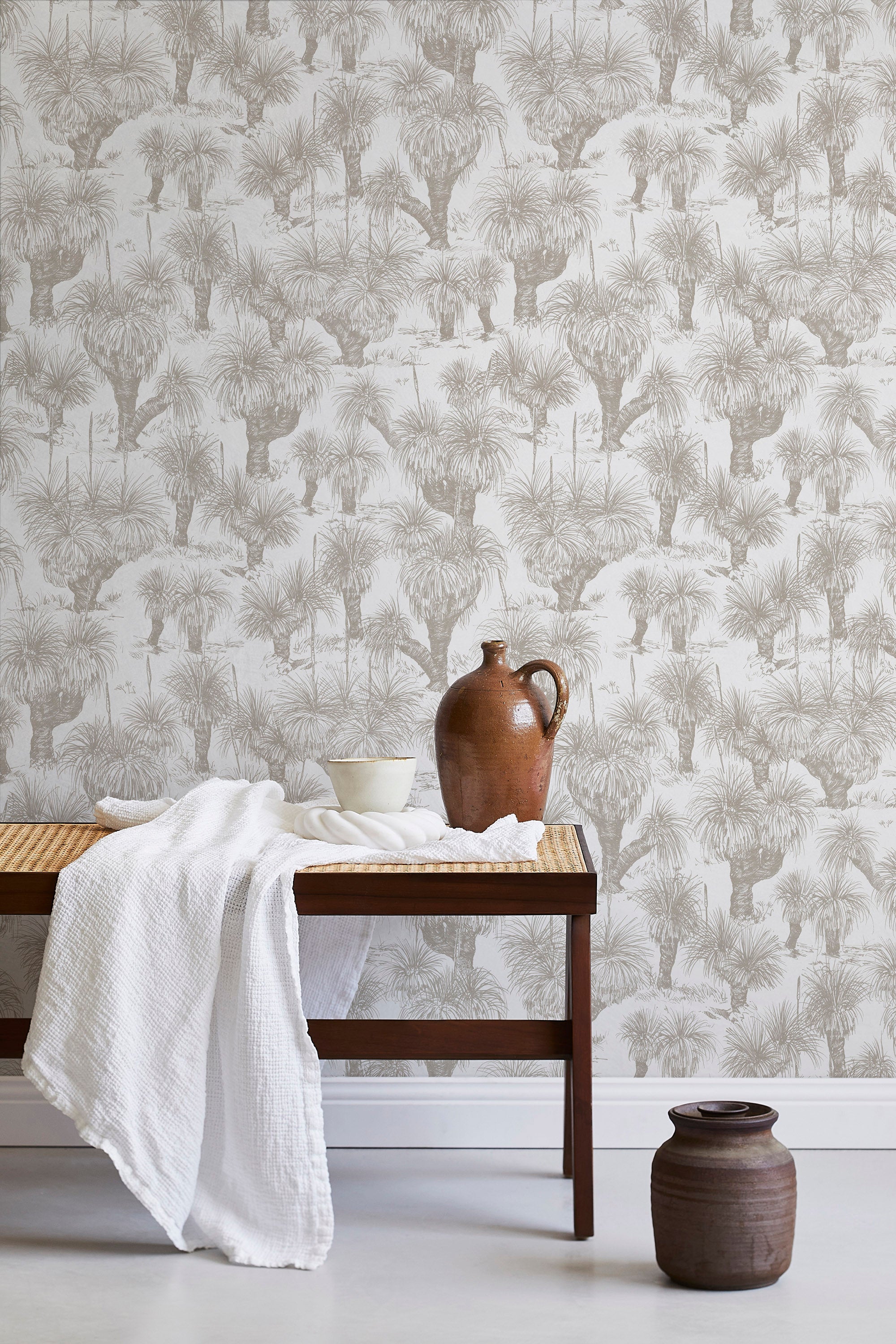 A bench with a towel and clay vases stands in front of a wall papered in a painterly tree print in tan and white.