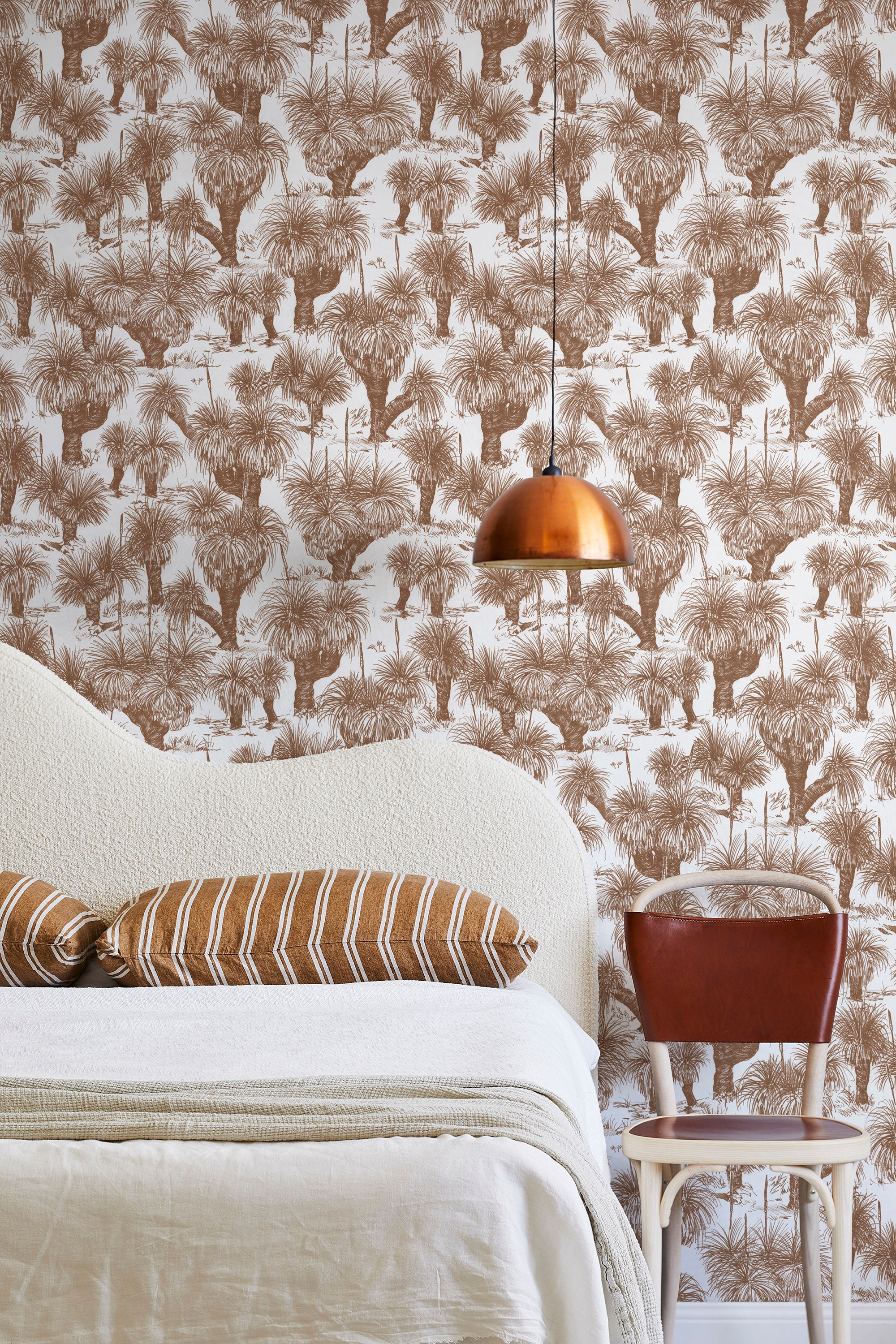 A modernist bed, hanging lamp and chair stand in front of a wall papered in a painterly tree print in rust and white.