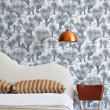 A modernist bed, hanging lamp and chair stand in front of a wall papered in a painterly tree print in navy and white.