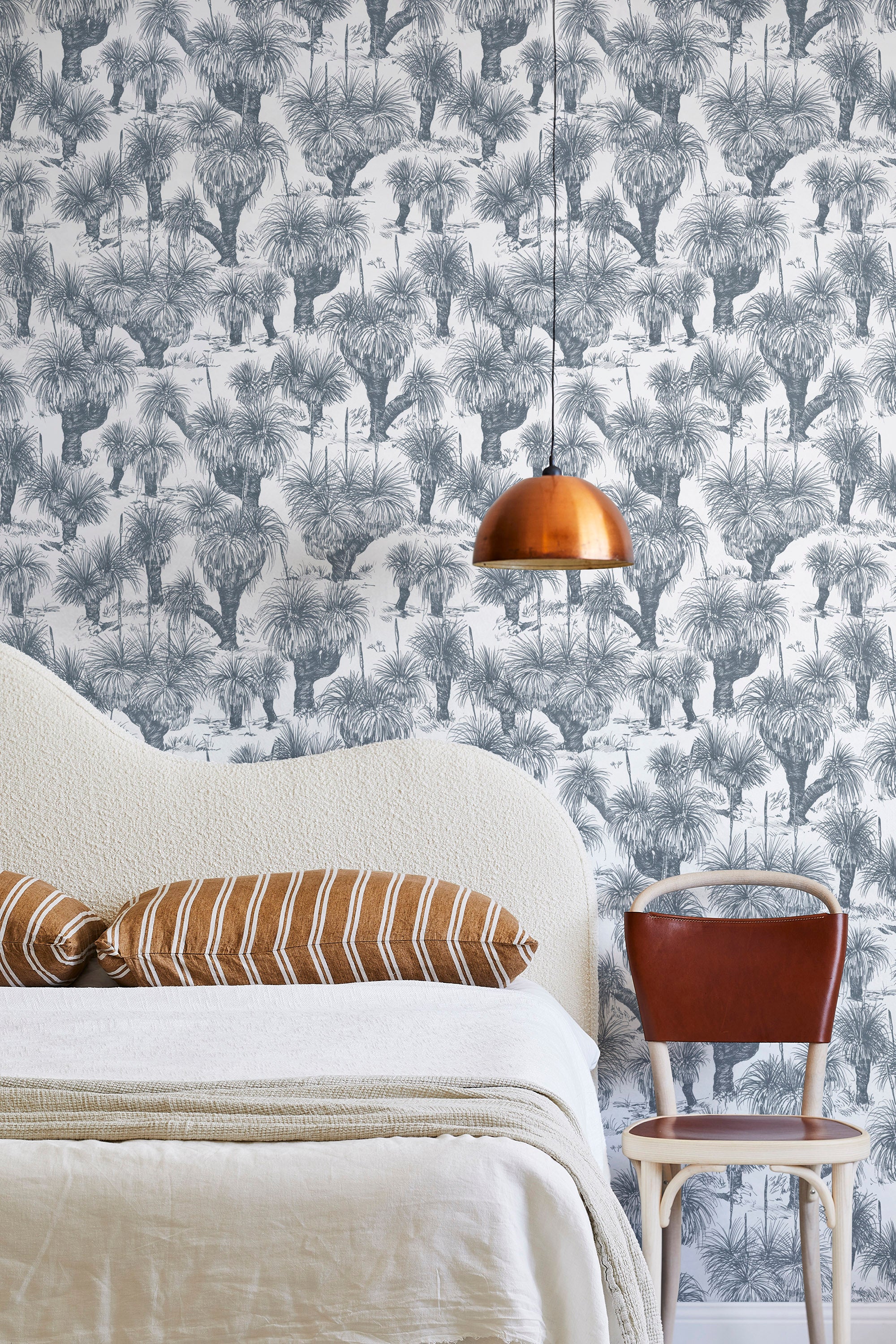 A modernist bed, hanging lamp and chair stand in front of a wall papered in a painterly tree print in navy and white.