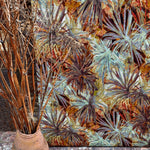 A vase of branches stands in front of a wall upholstered in a painterly palm tree print in shades of orange and brown.