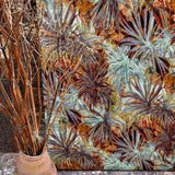 A vase of branches stands in front of a wall upholstered in a painterly palm tree print in shades of orange and brown.