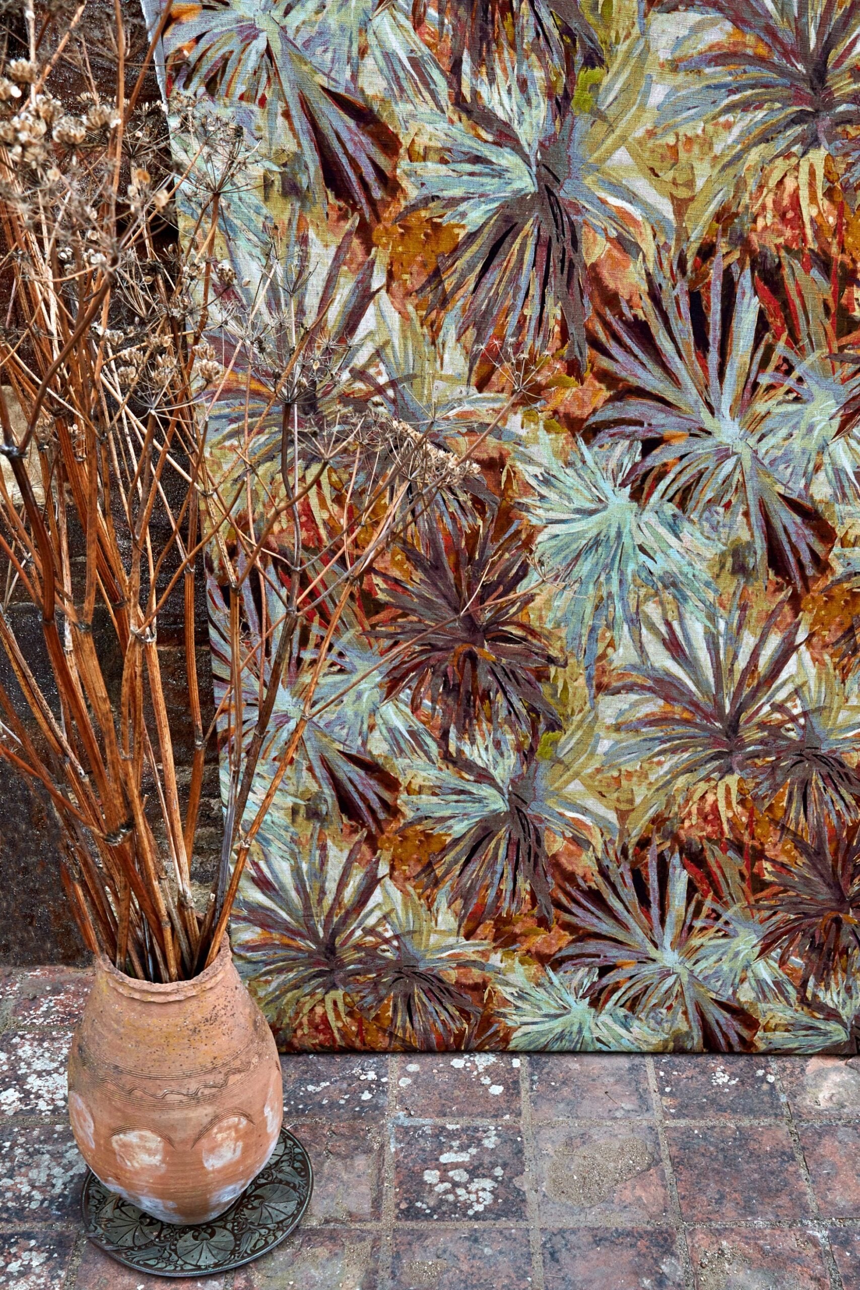 A vase of branches stands in front of a wall upholstered in a painterly palm tree print in shades of orange and brown.