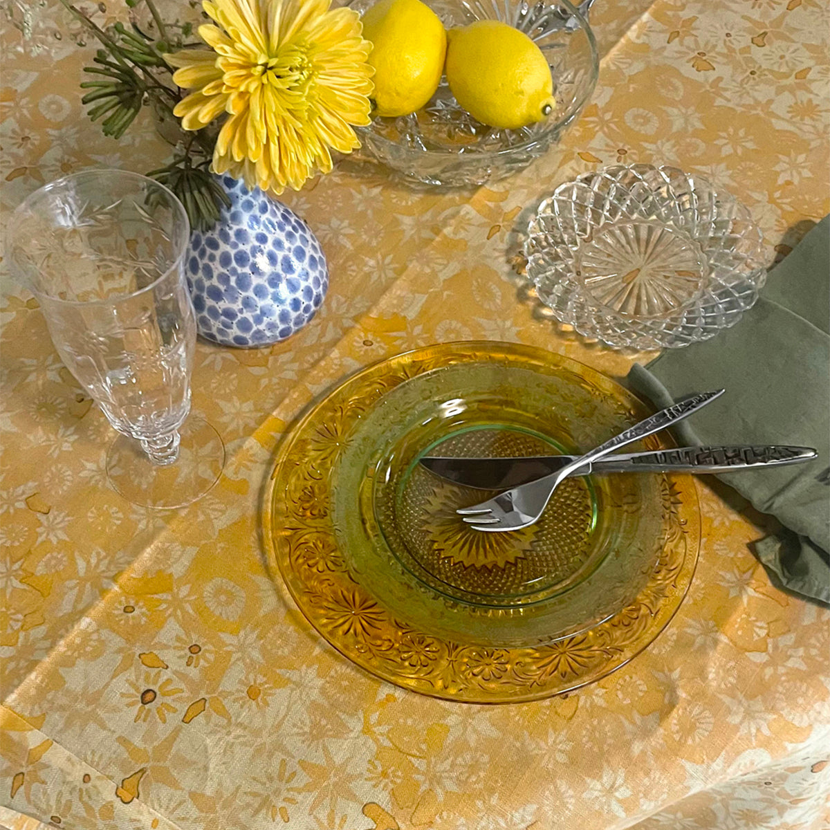Dining table setting with yellow flowers, lemons, and glassware on a patterned tablecloth.