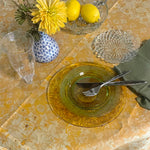 Dining table setting with yellow flowers, lemons, and glassware on a patterned tablecloth.