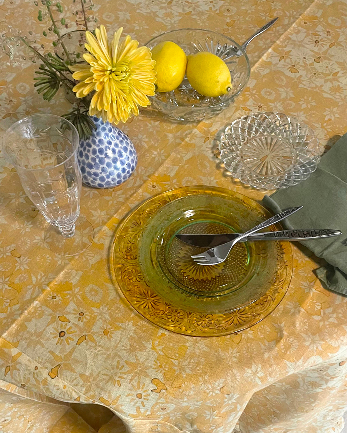 Dining table setting with yellow flowers, lemons, and glassware on a patterned tablecloth.