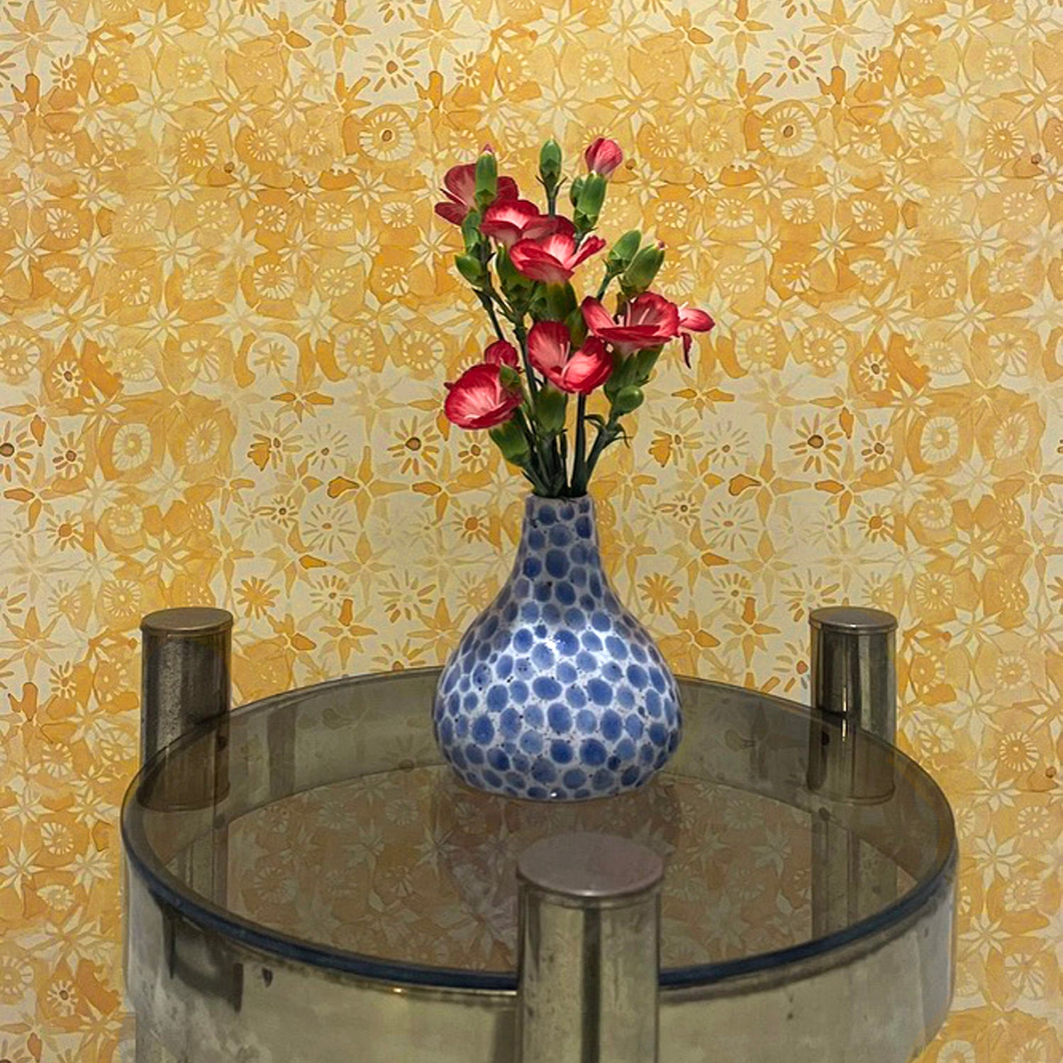 Small round table with a blue polka dot vase and pink flowers against a yellow floral wallpaper.