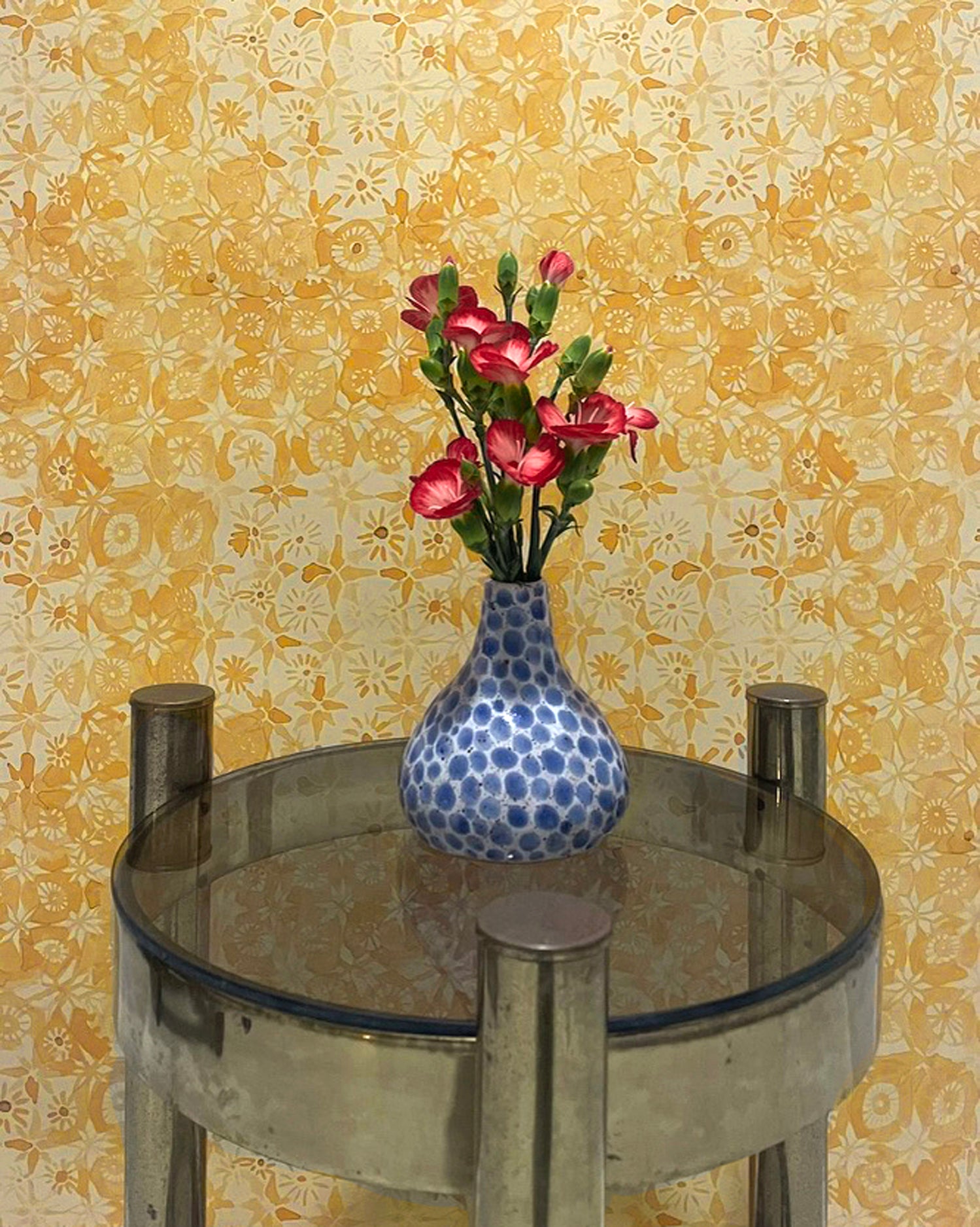 Small round table with a blue polka dot vase and pink flowers against a yellow floral wallpaper.