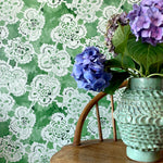 Decorative wallpaper with lace pattern, wooden stool, and floral arrangement.
